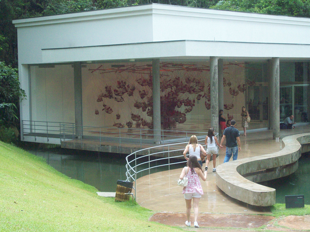 Vista à distância da obra True Rouge, de Tunga, uma instalação onde objetos de vidro pendurados possuem objetos e gosmas vermelhos dentro de si, parecendo grandes bolsas de sangue