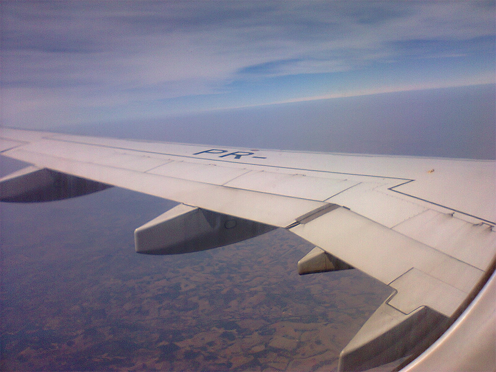 Foto de uma asa de avião em pleno voo, sobre as nuvens, tirada de dentro da aeronave.