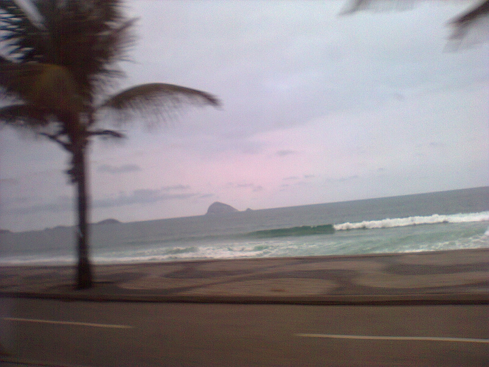 Foto de uma praia do Rio de Janeiro, tirada do outro lado da rua, sob céu muito nublado. O ambiente está vazia, em decorrência do tempo, e uma árvore que aparece à esquerda indica que estava ventando.