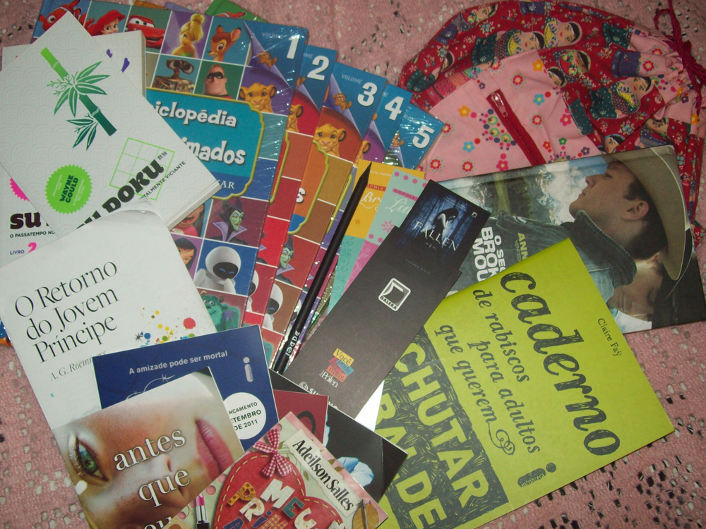 Fotografia tirada de cima mostrando os livros, brindes e lembranças trazidos da Bienal do Livro do Rio de 2011 espalhados sobre uma colcha rosa. Entre os itens aparecem exemplares de livros juvenis e romances, uma coleção da Enciclopédia Disney ainda lacrada, marcadores de página, folhetos, uma bolsa estampada com bonecas russas, uma revista com um cantor na capa e um exemplar do livro Caderno de rabiscos para adultos que querem chutar o balde.
