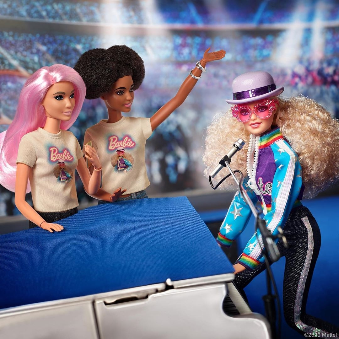 Barbie Elton John: imagem da boneca ao piano, tocando, com duas bonecas ao lado assistindo, uma branca de cabelos rosa e a outra negra de maria chiquinha. Ambas vestem uma camiseta com a imagem da boneca principal.