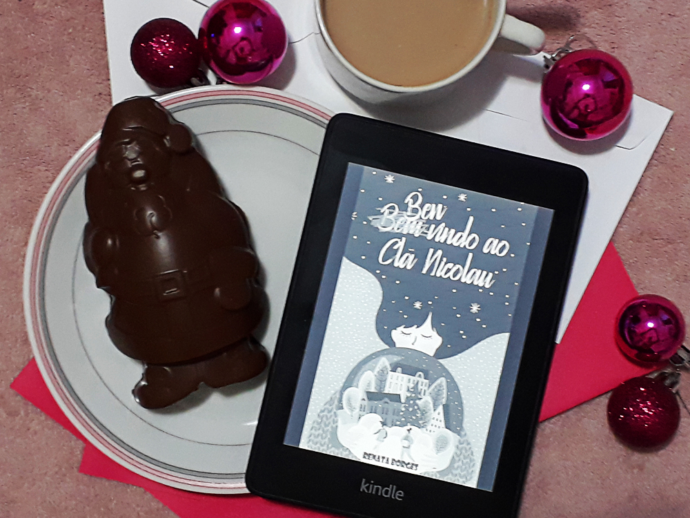 Bem-Vindo ao Clã Nicolau: foto do aparelho Kindle com a tela ligada onde a capa do livro aparece em destaque. Ao fundo, envelopes de papel brancos e cor-de-rosa, e ao redor bolas de natal rosa em tamanhos variados, uma xícara de chocolate quente e um prato contando um papai noel de chocolate.