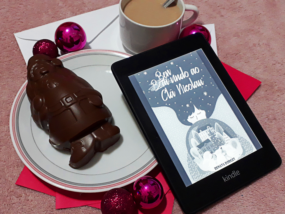 Bem-Vindo ao Clã Nicolau: foto do aparelho Kindle com a tela ligada onde a capa do livro aparece em destaque. Ao fundo, envelopes de papel brancos e cor-de-rosa, e ao redor bolas de natal rosa em tamanhos variados, uma xícara de chocolate quente e um prato contando um papai noel de chocolate.