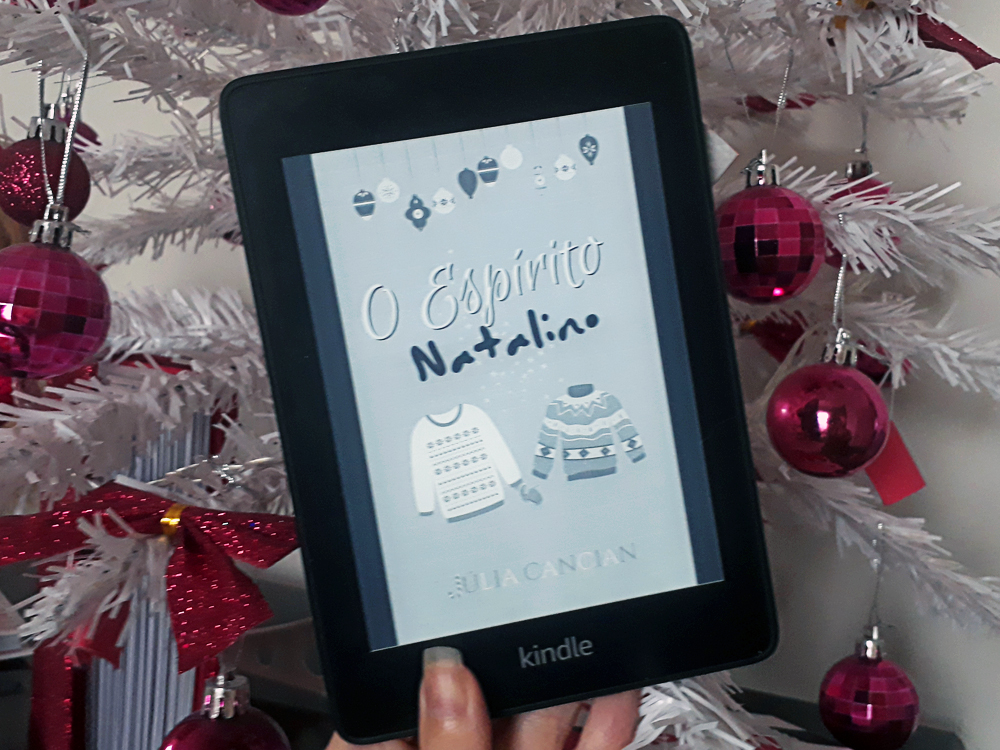 O Espírito Natalino: foto do aparelho Kindle com a tela ligada em que a capa do livro aparece em destaque. Ao fundo, galhos de uma árvore de natal branca com enfeites rosa choque variados.