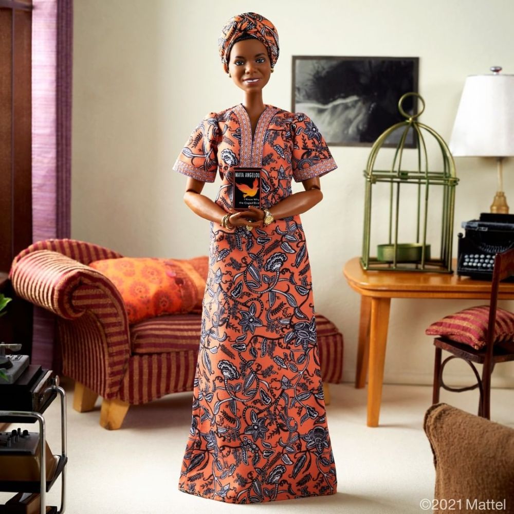 Barbie Maya Angelou: foto de corpo inteiro da boneca, que veste vestido e turbante alaranjados com a mesma estampa, de pé em uma sala em miniatura de aparência confortável.