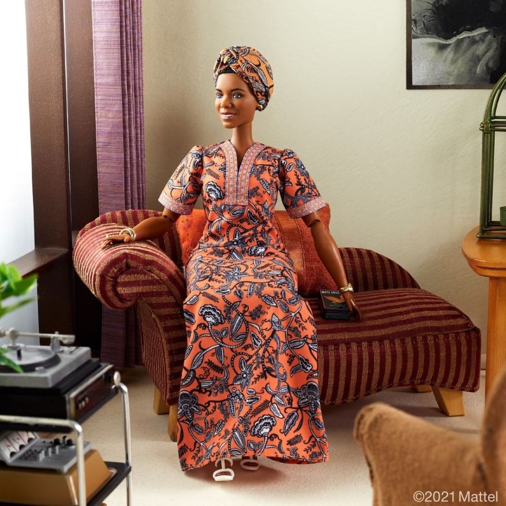 Barbie Maya Angelou: foto de corpo inteiro da boneca, que veste vestido e turbante alaranjados com a mesma estampa, sentada em uma sala em miniatura de aparência confortável.