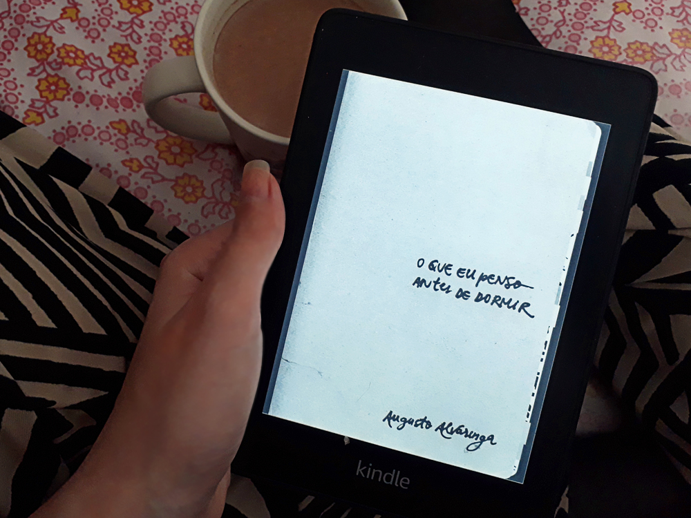 o que eu penso antes de dormir: mão de pele clara segurando um aparelho Kindle com a capa do livro na tela, lisa, em tom neutro, com o título ao centro alinhado à esquerda e nome do autor na lateral inferior esquerda. O e-book está apoiado em pernas cruzadas, usando calça de estampa geométrica, e ao fundo um tecido de tema floral e caneca contendo bebida achocolatada.