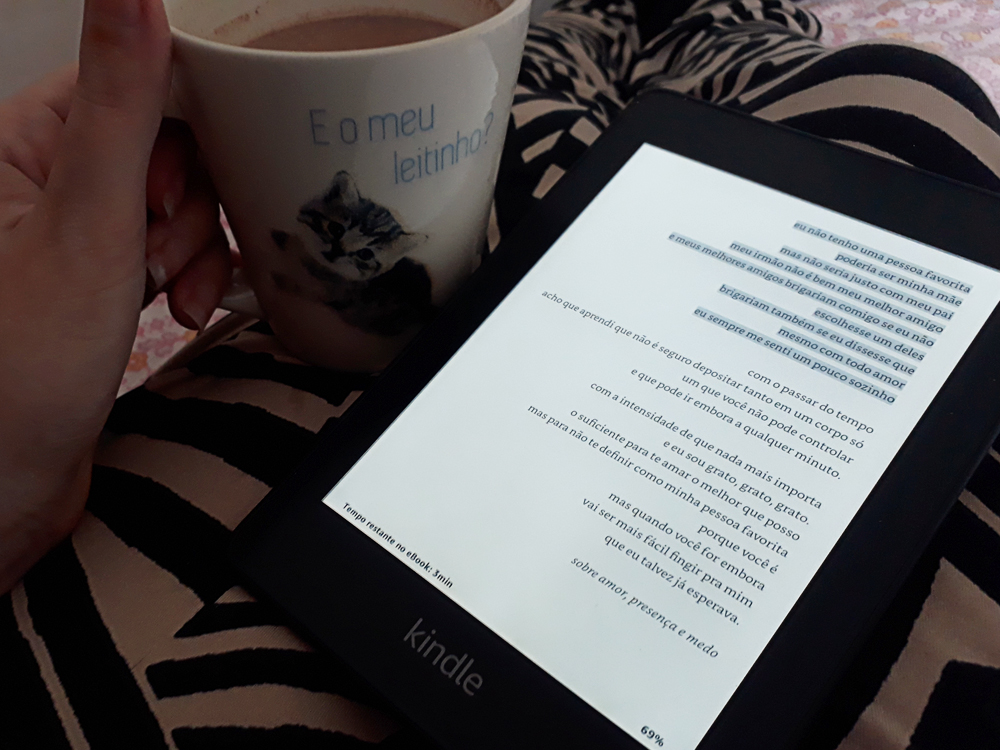 o que eu penso antes de dormir: mão de pele clara segurando uma caneca contendo bebida achocolatada atrás de um aparelho Kindle com uma página do livro na tela, o primeiro verso do poema está em destaque, marcado pela leitora no próprio aparelho. O e-book está apoiado em pernas cruzadas, usando calça de estampa geométrica, e ao fundo um tecido de tema floral.