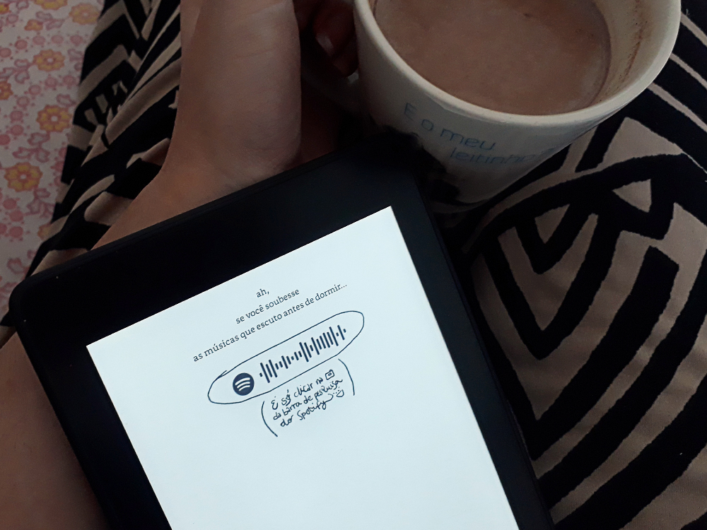 o que eu penso antes de dormir: mão de pele clara segurando uma caneca contendo bebida achocolatada atrás de um aparelho Kindle com uma página do livro na tela, contendo o código Spotify da playlist do mesmo. O e-book está apoiado em pernas cruzadas, usando calça de estampa geométrica, e ao fundo um tecido de tema floral.