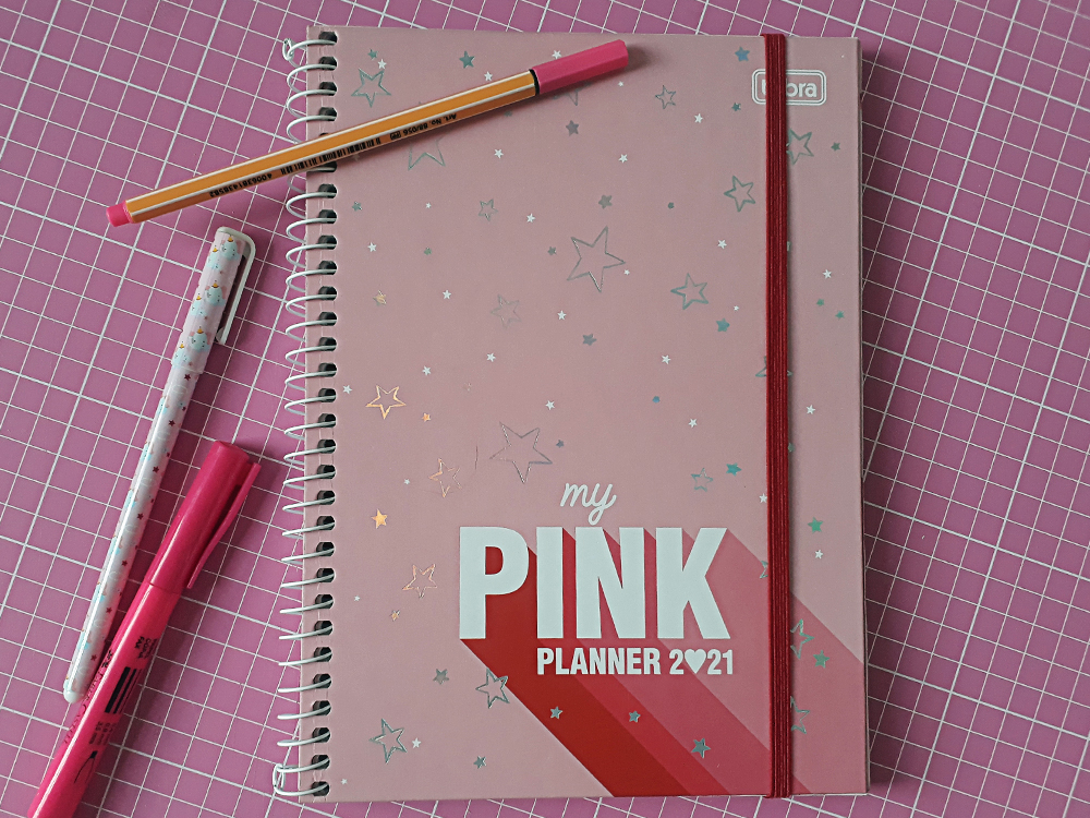 Capa do My Pink Planner Tilibra, rosa com detalhes em estrelas brilhantes, lateral em espiral e o nome do produto em baixo, escrito em branco. Ao fundo uma base de corte e materiais de escritório, todos também cor de rosa.