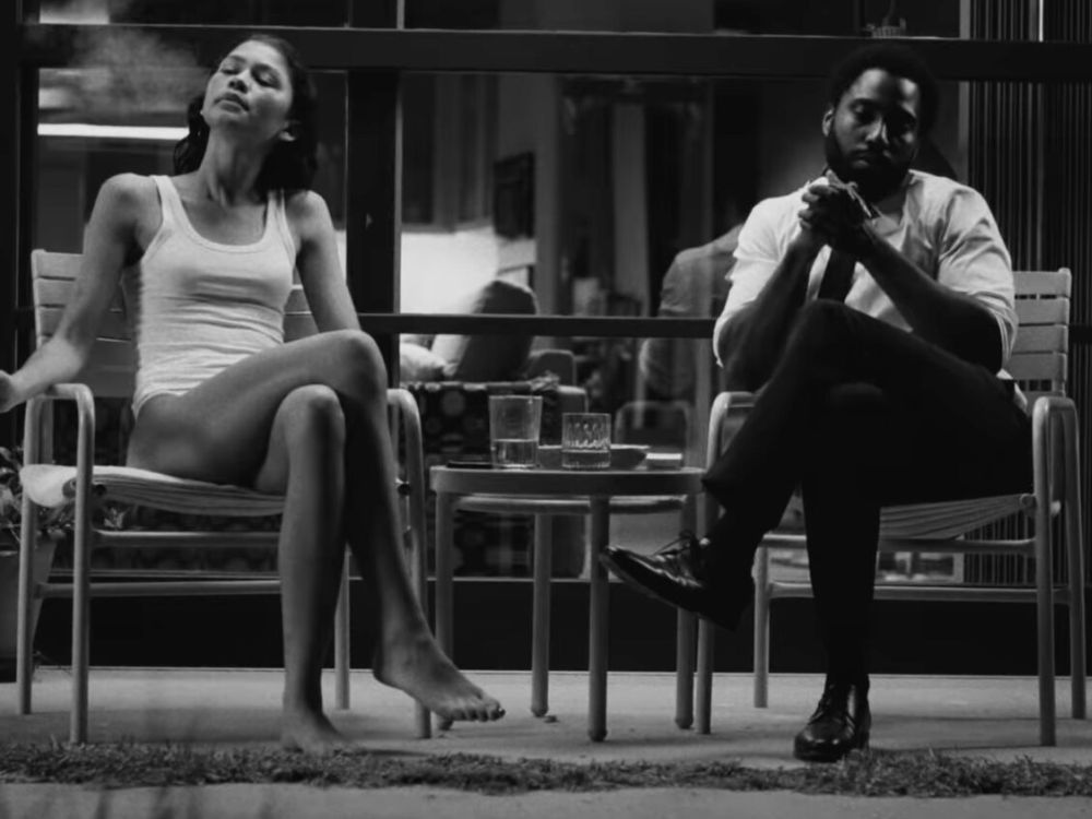 Malcolm & Marie: imagem em preto e branco do casal protagonista do filme onde ambos estão em um ambiente aberto, sentados em cadeiras de jardim com uma mesa entre eles. Marie veste apenas calcinha e uma camiseta e está soprando a fumaça do cigarro que está em sua mão, fora do enquadramento da imagem, enquanto Malcolm olho para suas mãos, que está juntas e levantadas em frente ao seu peito.
