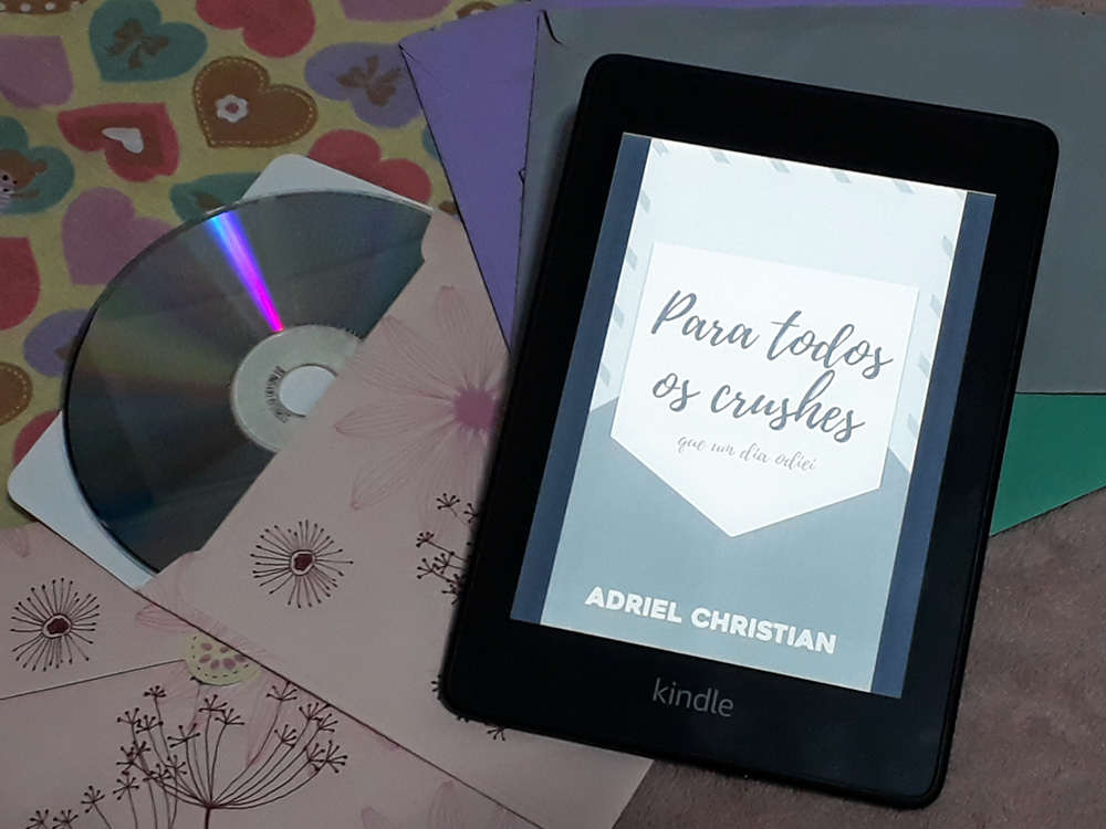 Para Todos os Crushes Que um Dia Odiei: aparelho Kindle ligado aberto na capa do livro onde se lê o título sobre formas geométricas que formam um envelope através do contraste de tons de cinza. Atrás, envelopes coloridos, tecidos com estampas de coração e um CD saindo de uma capa floral compõe o fundo da foto.