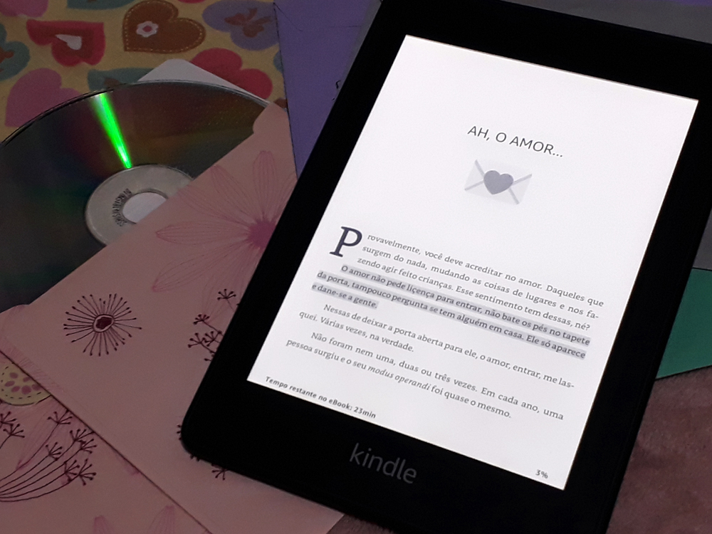 Para Todos os Crushes Que um Dia Odiei: aparelho Kindle ligado aberto em página do livro onde se lê o título 'Ah, o amor' e uma introdução do autor sobre a proposta do mesmo. Atrás, envelopes coloridos, tecidos com estampas de coração e um CD saindo de uma capa floral compõe o fundo da foto.