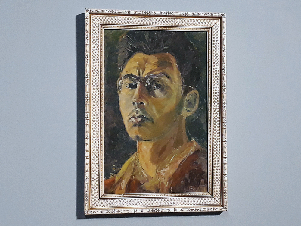 Autorretrato de Abraham Palatnik, que cuja imagem olha diretamente para o expectador com o rosto levemente virado. As cores do quadros se apresentam em grandes blocos disformes ao retratar um homem de pele clara, cabelos escuros e óculos de armação fina. Ele veste uma camisa em tons terrosos e a obra possui moldura de cor clara com detalhes ornamentações delicados.