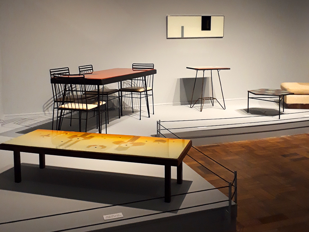 Móveis projetados por Abraham Palatnik: mesa de centro baixa, detangular, feita de madeira escura com tampo decorado em tons outonais formando objetos geométricos, mesa de jantar de metal com tampo colorido em tom forte acompanhada de quatro cadeiras de metal escuro com estofado claro, mesas de apoio de metal fino e escuro (uma alta, da altura da mesa de jantar, e a outra baixa, como a mesa de centro) com tampos decorados geometricamente, quadro minimalista de fundo claro com duas formas retangulares (uma escura a a outra, menos, em tom médio semelhante ao da moldura) e pequeno assento estofado em tom neutro, cortado pela moldura da foto.