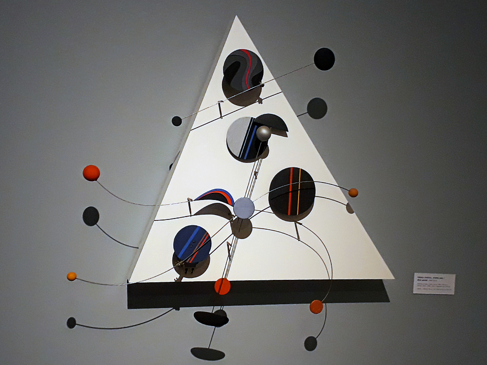 Objeto cinético de fundo triangular, com formas circulares planas e maciças coloridas fixadas em cabos retos e ondulares, permitindo movimento determinado pelo artista, não perceptível na imagem, que é estática.