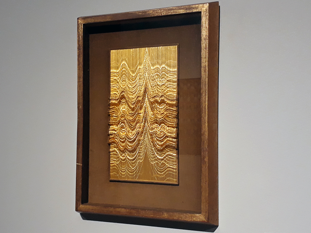 Quadro em metal dourado, cortado de forma que haja variações na cor do mesmo ao formar onda e picos pontudos. A obra está fixada em moldura de madeira com um vidro na  frente.