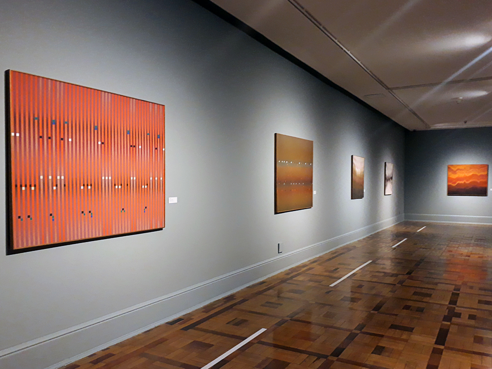 Sala da exposição da mostra Abraham Palatnik - A Reinvenção da Pintura com chão de taco e parede em tom neutro, onde se vê quatro quadros na parede lateral (três em tons quentes, um em cores neutras) e um na parede do fundo (cores quentes). No chão é possível ver marcações brancas em frente de cada obra, determinando a distância segura que o expectador deve ficar das mesmas.