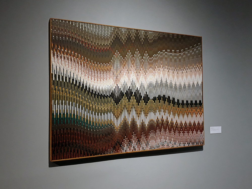 Quadro colorido, em diversas cores e tipos de tons, onde o uso de linhas de diferentes espessuras formando ondas e picos possibilitam efeitos de ilusão de ótica.