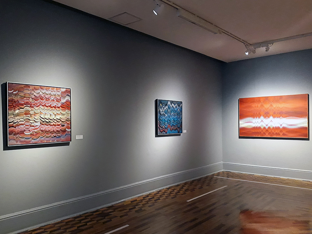 Sala da exposição com chão de taco e parede em tom neutro, onde se vê dois quadros na parede à esquerda (um em tons quentes e outra em tons frios) e um na parede da direita (tons quentes). As obras usam mistura de cor e uso de ondas e picos para criar efeito de ilusão de ótica. No chão é possível ver marcações brancas em frente de cada obra, determinando a distância segura que o expectador deve ficar das mesmas.