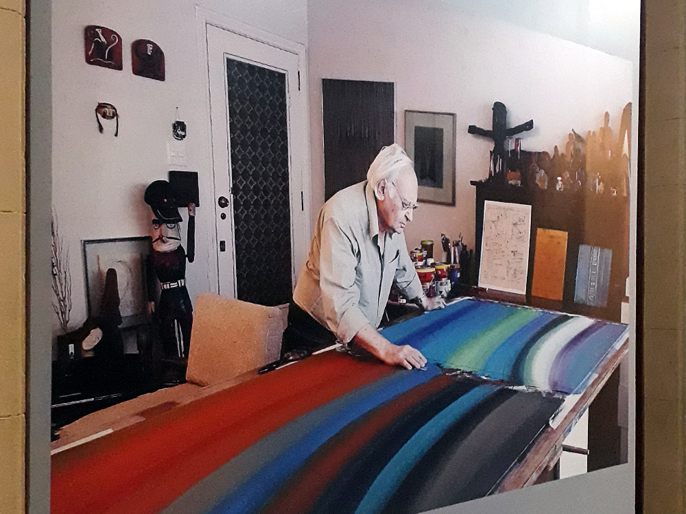 Foto de Abraham Palatnik em que o artista se olhando para um de suas obras, que está sobre uma mesa, um quadro feito de largas linhas de muitas cores diferentes, frias e quentes. Nela, ela aparenta ser idoso, com cabelos brancos e rosto enrugado, veste camisa de cor clara, calça preta e óculos de grau. Atrás dele é possível ver uma parte de seu ateliê, com quadros nas duas paredes brancas, alguns materiais artísticos (como tintas e pincéis) em uma mesa de apoio ao seu lado e uma porta de madeira com vidro ao fundo. Os m´veis, que também estão presentes em outra mesa de apoio e uma estante, possuem tons escuros de madeira.