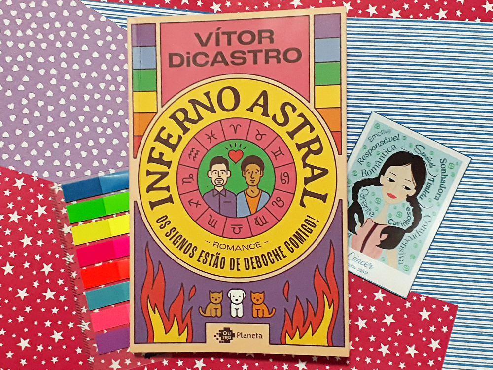 Inferno Astral: foto do livro sobre papéis de cores variadas de estampas diferentes (estrelas, corações e listras). Embaixo dele, ainda sobre o papel, há um conjunto de flags para marcar livros de várias cores e um ímã de geladeira com uma ilustração remetente ao signo de Câncer, onda a menina tem as pontas das tranças em forma de pinças de caranguejo e qualidades do signo escritas ao seu redor.