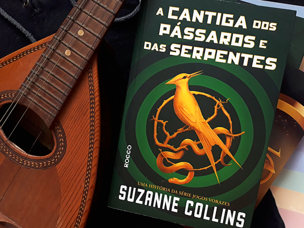 Instrumento de cordas pequeno e marrom à esquerda da imagem, ao lado do livro resenhado. Ao fundo, uma camiseta preta com o símbolo da saga Jogos Vorazes, um pedaço de papel arco-íris em um canto e um marador de livro da saga Jogos Vorazes ao lado.