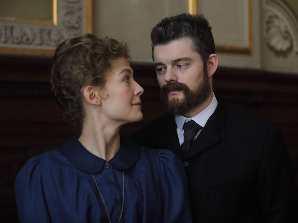 Radioactive: Rosamund Pike e Sam Riley, interpretando o casal Curie, parados vestindo roupas escuras lado a lado, ela levemente na frente, se olhando.