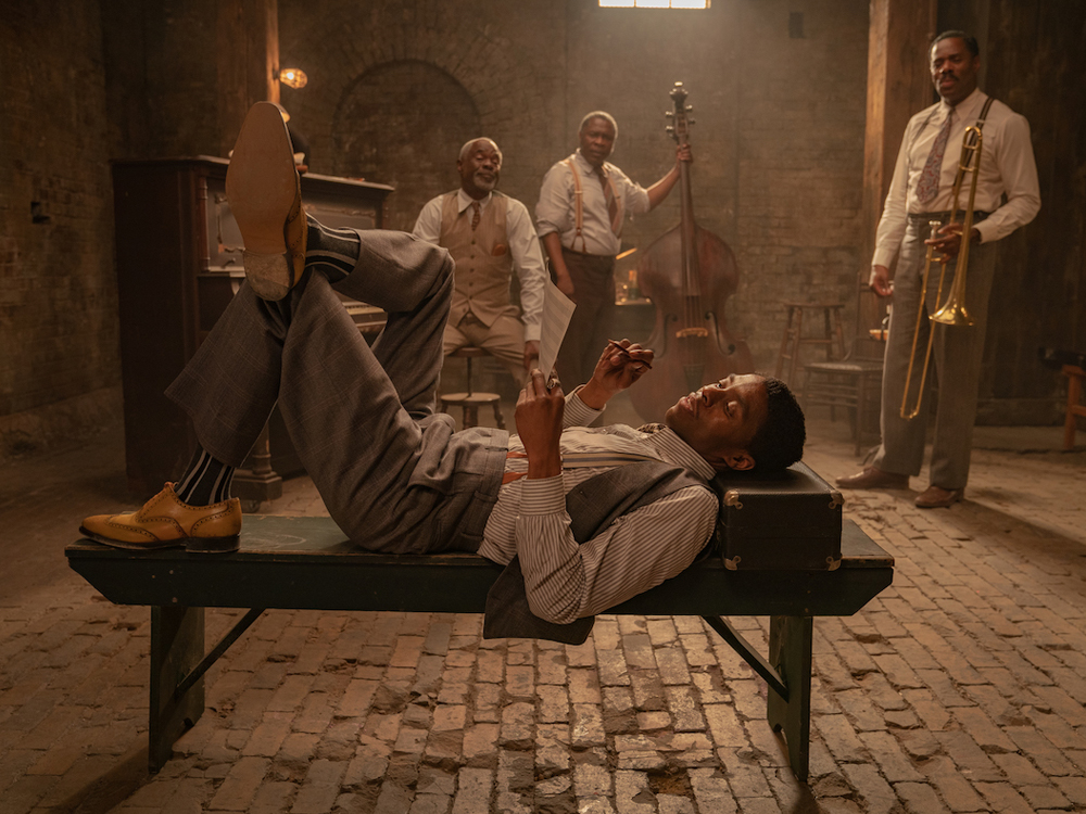 A Voz suprema do Blues: cena do filme em que Chadwick Boseman se encontra em primeiro plano, deitado sobre um banco com a cabeça apoiada em uma melta, e os outros membros da banda do filme estão ao fundo, olhando pra ele, ao lado de seus instrumentos. O quarteto se encontra em uma sala de parede de tijolos expostos e piso de paralelepípedos, com apenas uma pequena janela ao fundo de onde vem a iluminação e toda o ambiente tem tons neutros evidentes, dando detaque para o dourado dos instrumentos de sopro e o amarelo do sapato do protagonista.