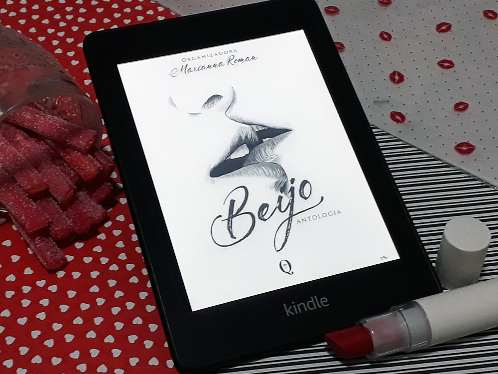 Aparelho Kindle ligado na capa do livro Beijo, de vários autores. A capa é toda branca, com o sombreado de duas bocas se beijando e o título em letra refinada em cima. Acima está o nome da organizadora e abaixo a logo da editora. A foto também contém um batom vermelho de embalagem branca abaixo e um copo deitado com balas de gelatina compridas também vermelhas dentro à direita. O fundo é composto de papéis de três texturas diferentes: vermelha com corações brancos, listrado de preto e branco e branco com bolinhas pretas e marcas de bocas vermelhas.