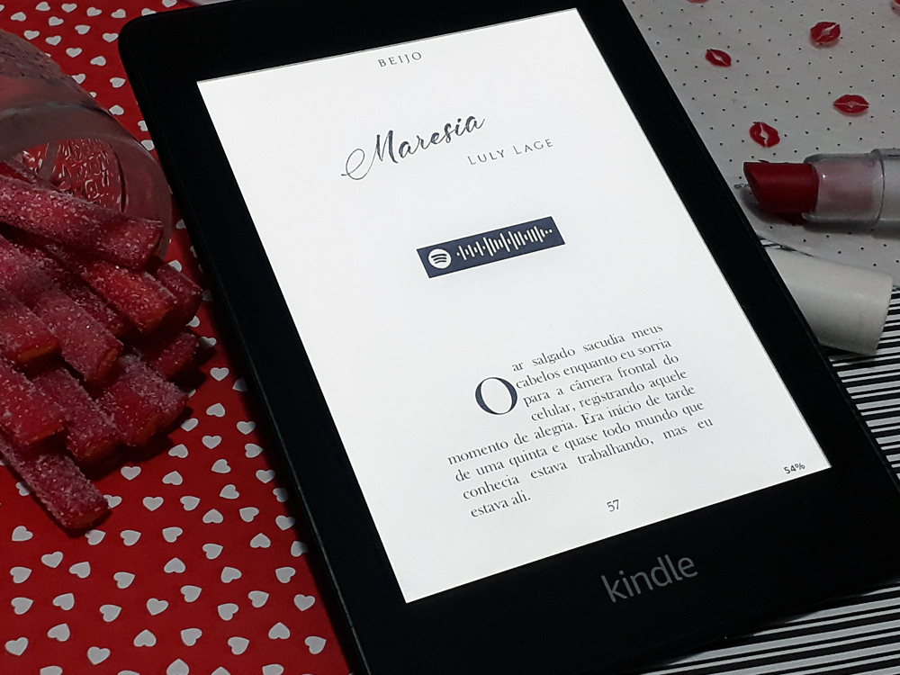 Aparelho Kindle ligado na página 57 do livro, primeira da crônica Maresia, de Luly Lage. Abaixo do título e do nome da autora há um código para ouvir a música tema no aplicativo Spotify e, em seguida, a história começa. O aparelho está inserido no mesmo cenário das outras fotos.