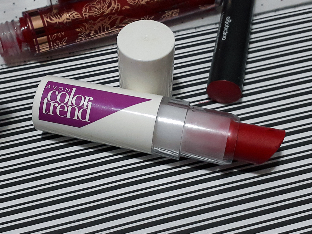 Batom Pimenta Matte, da marca Avon, linha Color Trend, aberto deitado sobre a superfície. Ao seu lado, partes dos outros batons estão visíveis também deitados.