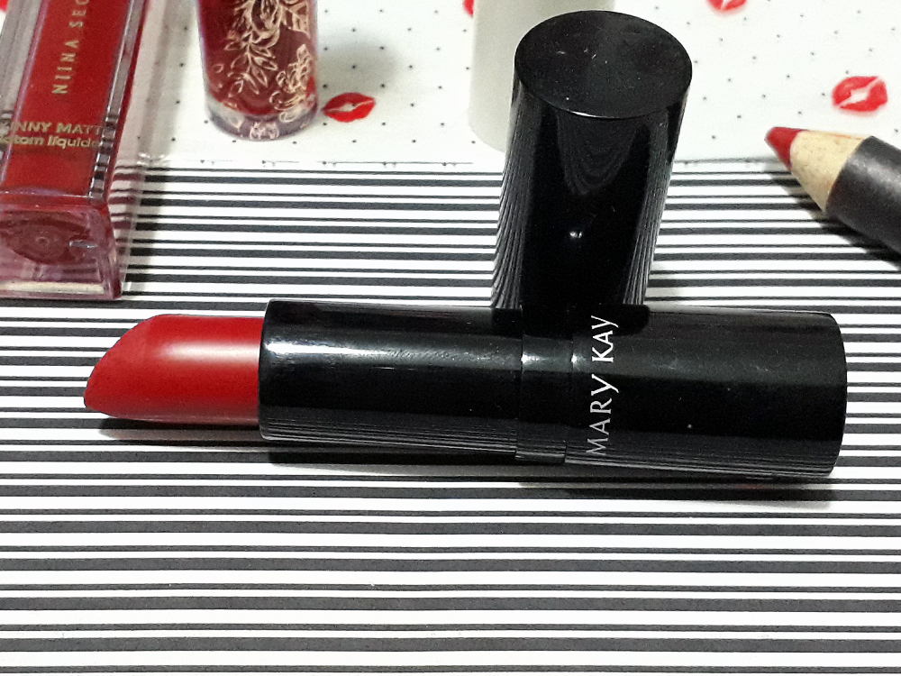 Batom Roma Red, aberto, com a tampa ao lado, deitado, com partes dos outros batons citados no post colocados ao seu redor, aparecendo parcialmente nas laterais da foto.