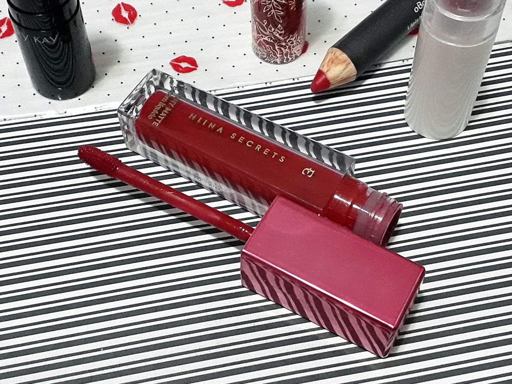 Batom líquido Vermelho Hibisco da Linha Niina Secrets para Eudora, com embalada quadricular e tampa rosa. O produto está aberto, deitado, com a tampa e aplicado ao lado.