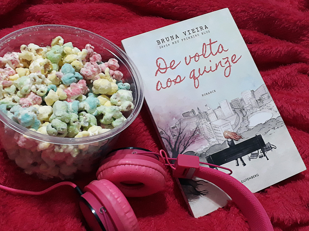 Balde de pipocas doces doloridas à esquerda, ao lado do livro De Volta aos Quinze fechado com a capa frontal virada para cima. Em baixo, parcialmente sobre o livro, há um fona de ouvido no modelo headphone rosa, e todos os elementos estão sobre uma coberta propositalmente levemente bagunçada rosa também.