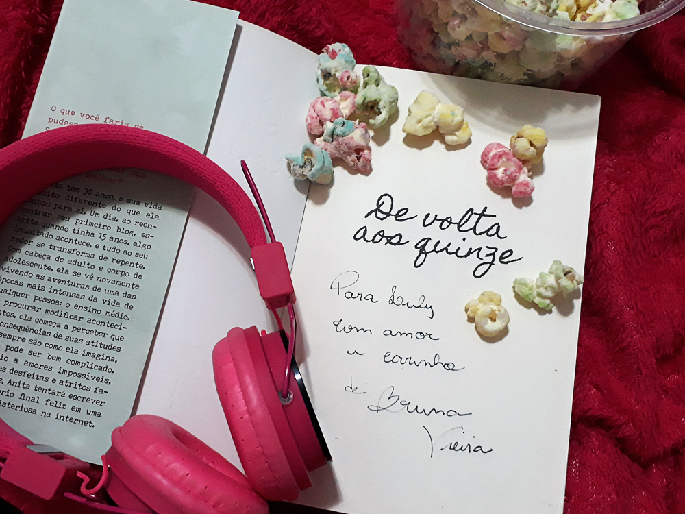 Livro De Volta Aos Quinze aberto na folha de rosto, onde está impresso seu título. Abaixo, à mão, há uma dedicatória com autógrafo da autora que diz 'Para Luly com amor e carinho de Bruna Vieira'. Existem alguma pipocas coloridas jgadas sobre essa página, com o balde cheio delas ao lado aparecendo parcialmente no topo da foto, e os headphones cor de rosa também estão ali, sobre a folha e a capa aberta do livro.