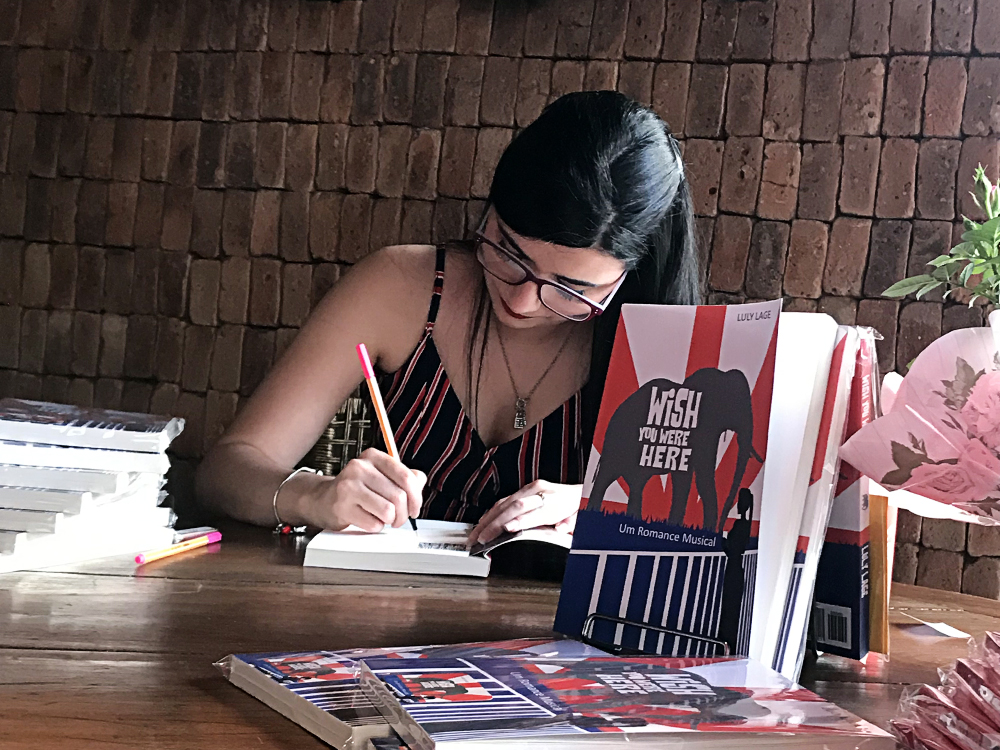 Foto de uma mulher de pele branca e cabelos escuro curvada sobre um livro enquanto o autografa usando uma caneta rosa. Ao seu redor, na mesa, várias edições do mesmo livro, Wish You Were Here: um romance musical, que tem capa da mesma cor da roupa que ela veste com uma menina segurando uma carta enquanto olha para um elefante ao fundo.