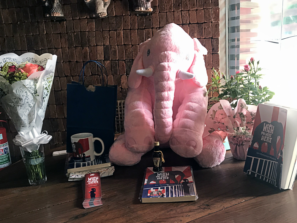 Mesa de madeira com alguns exemplares do livro, marcadores, caneca com reprodução da capa, um elefante de pelúcia rosa grande, uma sacola de presente azul, um buquê e um vaso de flores.