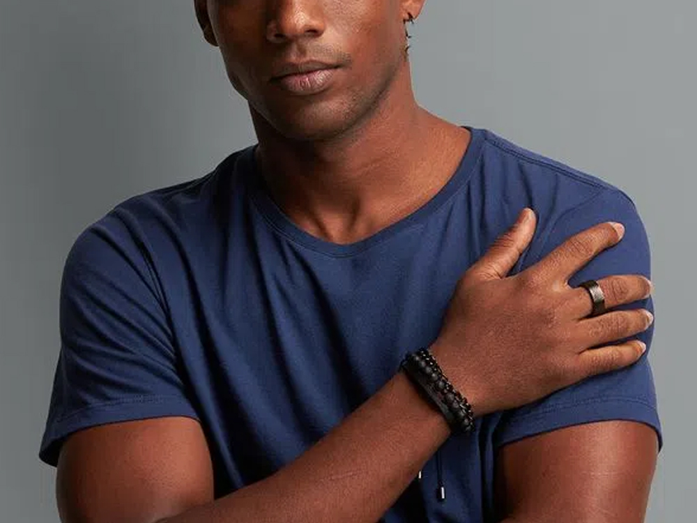 Foto de um homem negro, que mostra do nariz ao peito, usando uma camisa de gola careca azul. Sua mão direita está na frente do corpo, tocando o ombro esquerdo, e ele usa pulseiras artesanais.