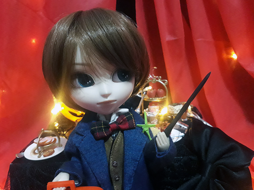 Foto do boneco vestindo como o personagem Newt Scamander, do filme Animais Fantásticos e Onde Habitam, usando camisa branca, gravata borboleta xadrez, colete marrom e casaco longo azul marinho, com uma miniatura da criatura Tronquilho, um humanoide feito de graveto de madeira verde, no ombro esquerdo. A mão esquerda segura uma varinha no ar e a direita uma maleta. Ao funo, se vê uma decoração em tons escuros, com algumas luzes pontuais, e uma mesa cheia de miniaturas de guloseiras.