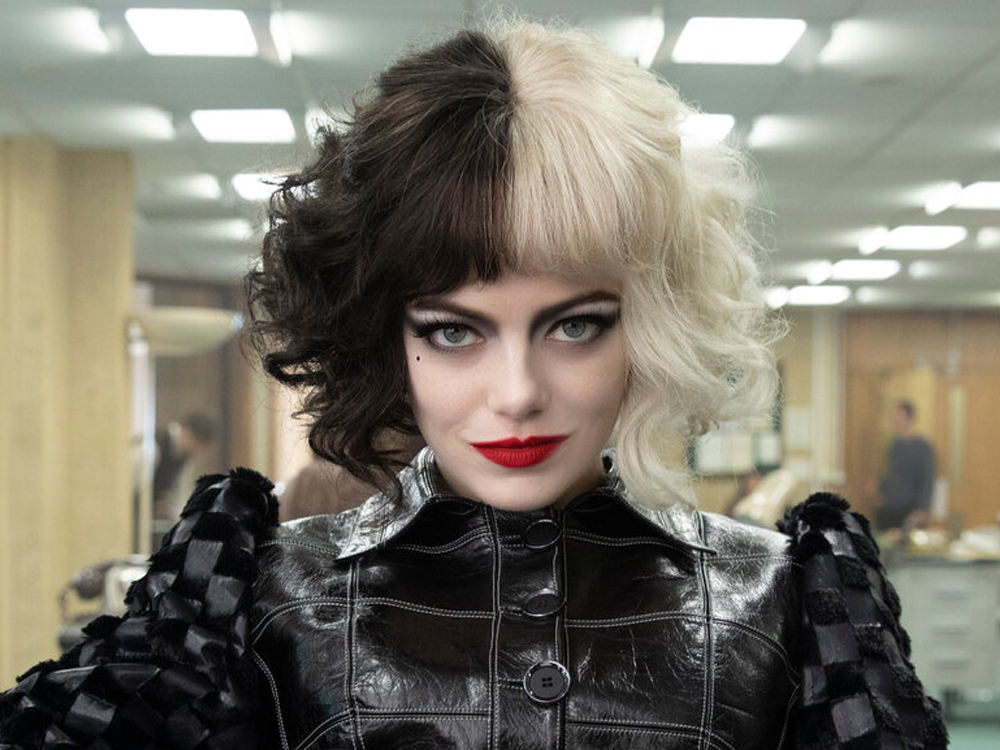 Foto da personagem Cruella olhando diretamente para a câmera com seus olhos azuis e olhar dissimulado. Ela está em um ambiente que parece uma redação de revista ou jornal, bem iluminado, e o close mostra apenas a parte de cima da sua roupa de couro preto pesada, os cabelos curtos metade da cor preta e a outra metade branca e o rosto, onde usa um batom vermelho vibrante.