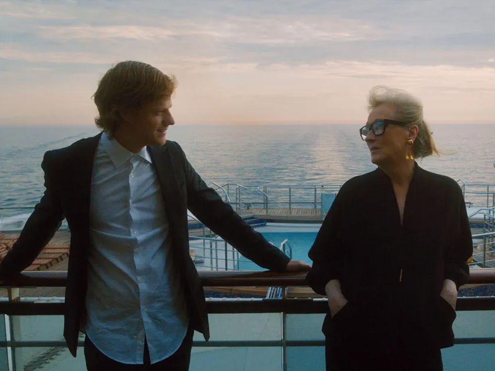 Foto de uma cena de Let Them All Talk onde as personagens Tyler e Alice, ele usando terno escuro e camisa de cor clara enquanto ela usa uma roupa completamente escura, encostados no parapeito de uma varanda do navio em que estão viajando. Os dois estão lado a lado, de frente para a câmera, olhando um para o outro, e ao fundo é possível ver um pouco do andar debaixo do navio e o oceano à sua frente.