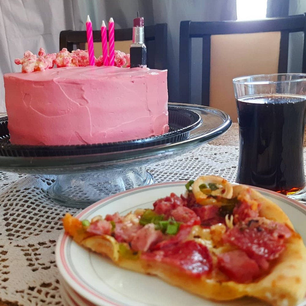 Foto do mesmo bolo presente na primeira imagem sobre o pequeno pedestal porta bolo de vidro. À sua frente um prato branco de bordas rosa contendo um pedaço de pizza de calabresa com alho poró e, à direita, entre os dois elementos, um copo de vidro cheio de Coca-Cola. Todos estão sobre uma mesa coberta por uma toalha branca.
