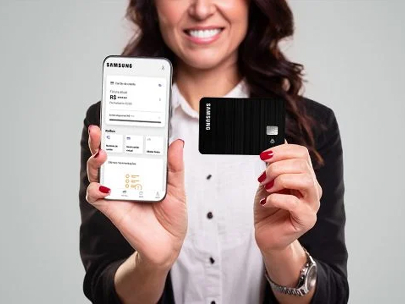 Imagem de uma mulher de pele clara e cabelos escuros sorrindo, segurando em uma mão um celular e na outra um cartão de crédito, ambos na direção do expectador.