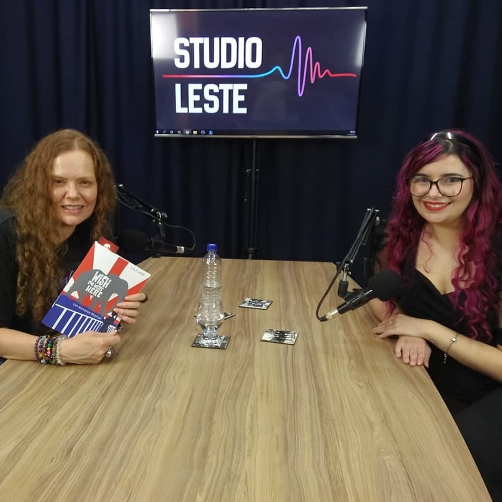 Foto de uma sala de paredes escura com uma televisão ao fundo onde se lê Studio Leste e uma mesa de madeira clara em frente com dois microfones em cima. Em lados aoposto, em frente a cada microfone, estão duas mulheres. Aninha Paixão veste uma blusa escura e tem cabelos compridos, ondulados e claros, além de um relógio no braço direito enquanto segura o livro Wish You Were Here: um romance musical, de Luly Lage. A autora está do outro lado, usando também uma roupa escura, cabelos coloridos cacheados e óculos de armação grossa.