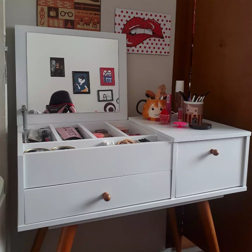 Foto de uma penteadeira branca com quatro pés palito em cor de madeira clara, com duas gavetas e uma tampa aberta, onde há um espelho fixado. Na parte interior de onde a tampa ficaria estão quatro divisórias com produtos de maquiagem dentro e na parte superior que não está levanta, sobre uma das gavetas, há duas canecas com pinceis de maquiagem ao lado de alguns produtos espalhados. Na parede existem dois quadros, um com ícones da história de Harry Potter e outro com uma boca vermelha mordendo o lábio inferior.
