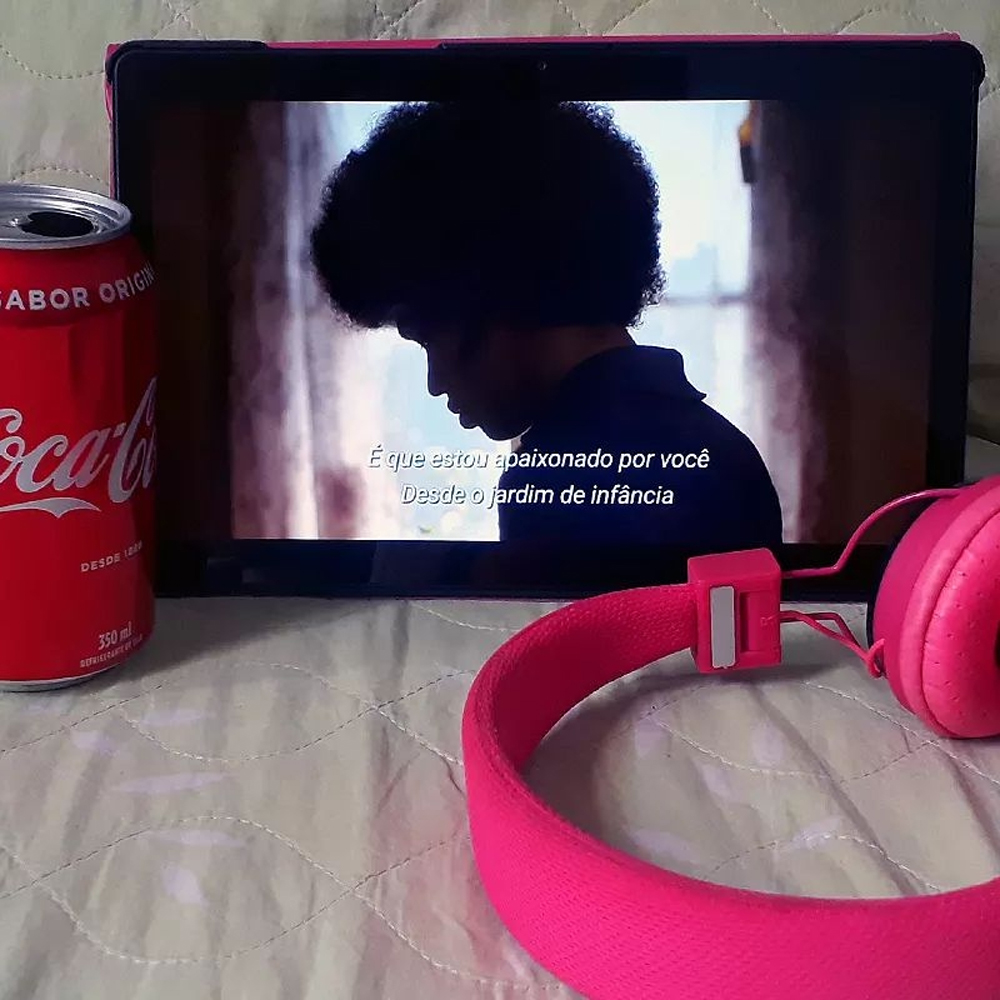 Foto de um tablet com uma cena da série The Get Down, onde o protagonista Zeke, um jovem negro de cabelos black power, está de lado, olhando para baixo. Abaixo há uma legenda onde diz 'Porque sou apaixonado por você desde o jordim de infância'. De um lado há uma lata do refrigerante vermelho Coca Cola à esquerda e um fone de ouvido rosa choque à direita.