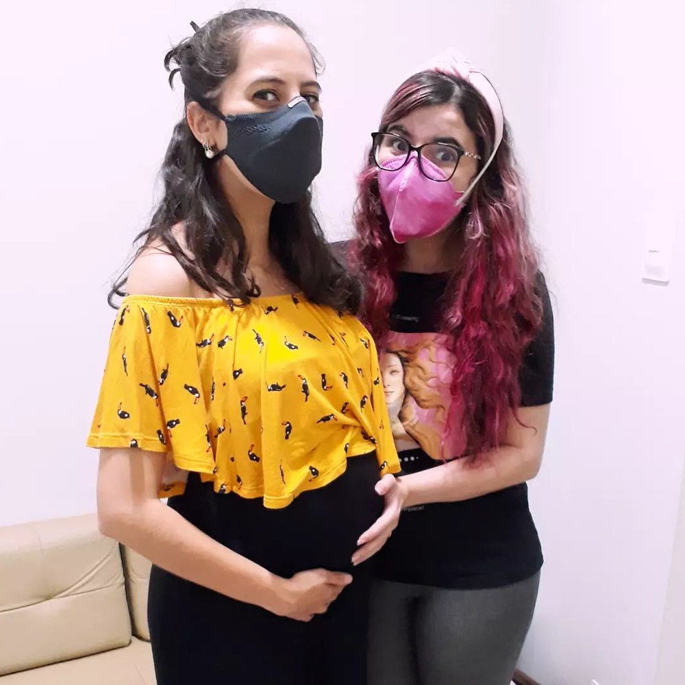 Foto de Luly Lage (mulher branca de cabelos compridos e rosa, vestindo camiseta preta, calça jeans e máscara de proteção rosa) segurando a barriga de grávida uma amiga que está ao seu lado (mulher branca, levemente mais alta, de cabelos escuros vestindo blusa amarela, calça preta e máscara de proteção preta).