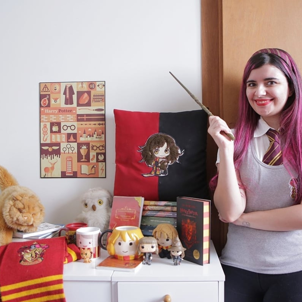 Foto de Luly Lage vestindo uniforme da Casa Grifinória dos filmes de Harry Potter enquanto empunha uma varinha apontada para cima e, ao seu lado, vários objetos referentes à saga, como alfomada, miniaturas, caneca (todos da personagem Hermione), pelúcias de gato e coruja, livros, etc. 