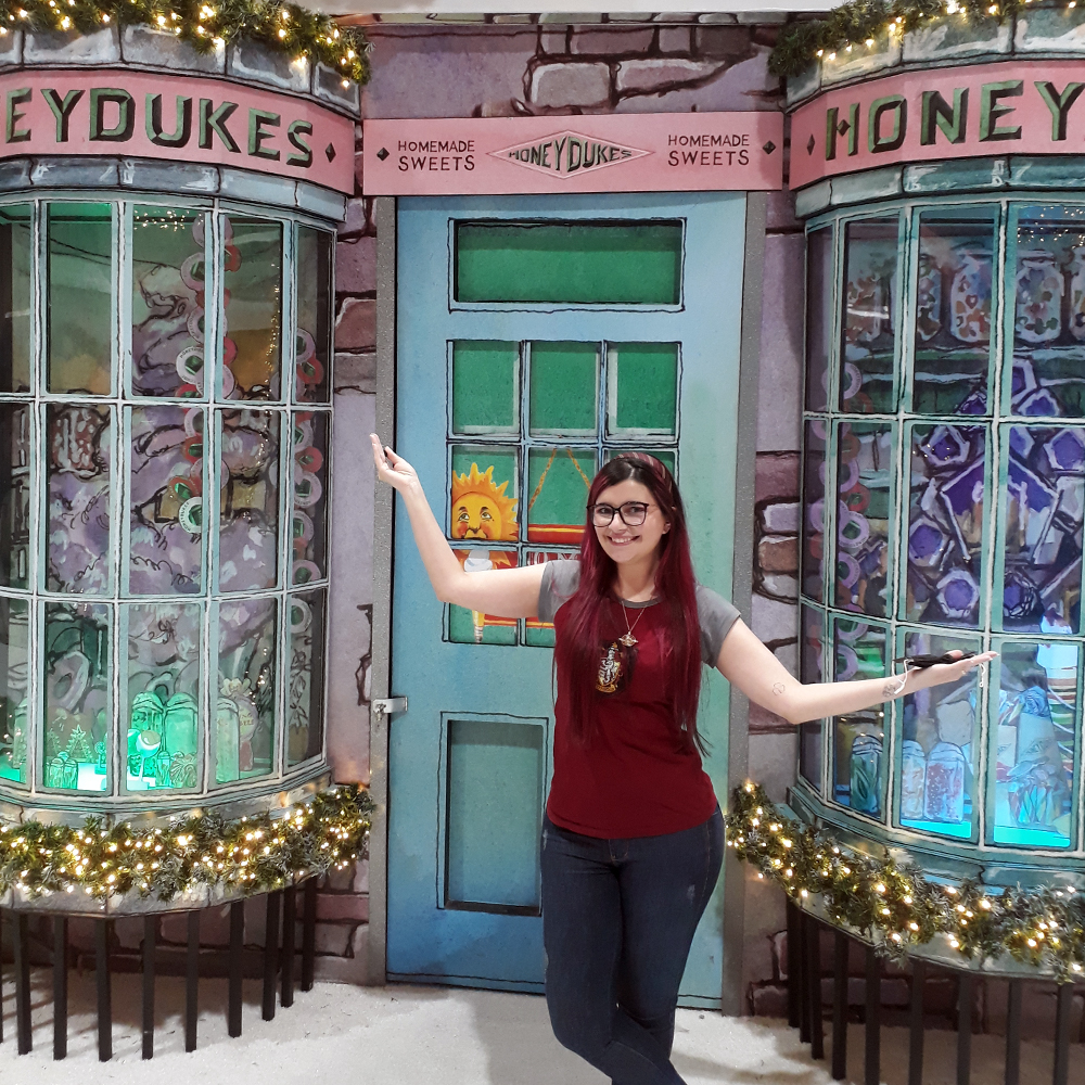 Foto de Luly Lage de pé, com os braços abertos recepcionando o expectador, em frente a uma simulação da loja Dedos de Mel do universo de Harry Potter, cuja fachada é colorida em tons pastéis e com a logo da loja ao topo.